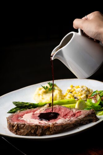 Sauce being poured over sliced beef with asparagus, corn, and mashed potatoes.