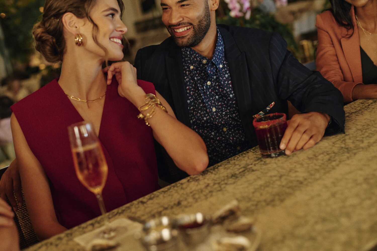 man and a woman sitting at the bar with cocktails looking at each other and smiling