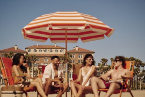 Four friends in swimsuits lounge under a beach umbrella, a luxury hotel in the background.