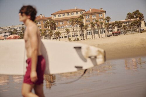 man holding a surfer board on the beach, out of focus, with Casa del Mar Hotel in the background
