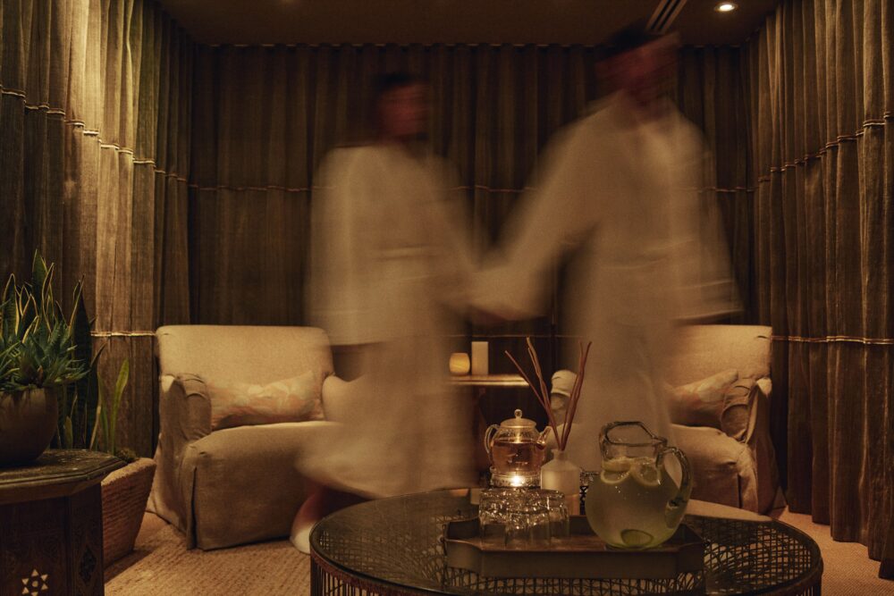 A man and woman in robes walk out of the frame in the hotel spa's sitting room, out of focus.
