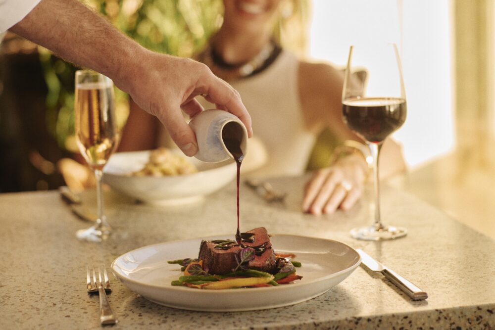 server pouring sauce on a plate of steak at a dinner table