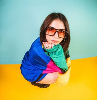 A woman with dark hair wears a bright, colorful jacket and large sunglasses.