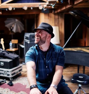 A man in a black hat and blue plaid shirt sits in a music studio, immersed in creativity.