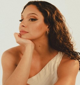 A curly-haired woman in a one-shoulder top gazes thoughtfully into the distance.