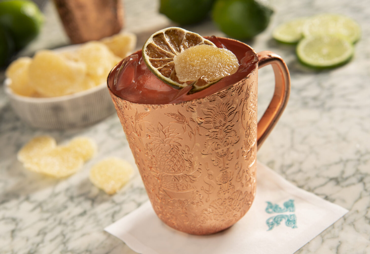 Copper mug with a drink garnished with lime and candied ginger, surrounded by lime slices.