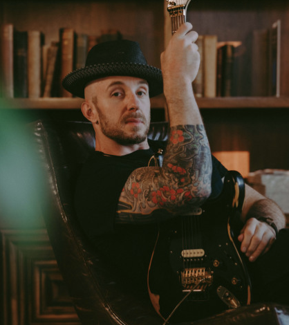 Chris Dalton with tattoos and a hat sitting, holding an electric guitar in a cozy room.