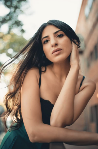 A woman with long dark hair in a black dress, posing outdoors with a gentle smile.