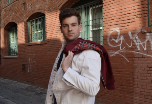 A man in a white jacket and red plaid scarf standing against a brick wall with graffiti.